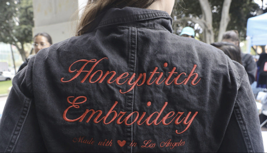 Black denim jacket with 'Honeystitch Embroidery' text and Denim Day logo on a blurred outdoor background.